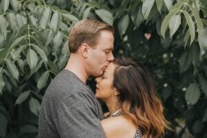 A couple kisses in front of green leaves during their engagement session.